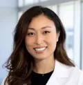 A woman with long, wavy brown hair wearing a white lab coat is smiling at the camera. She stands in a professional environment with large windows in the background, embodying the confidence and warmth you'd expect from one of the best urologists in Los Angeles.