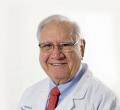 A smiling elderly man with white hair and glasses is wearing a white medical coat over a light blue shirt and an orange patterned tie. He proudly represents one of the best urologists in Los Angeles as part of our team at Comprehensive Urology. The background is plain white.