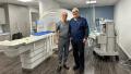 Two men in medical scrubs stand smiling next to each other in a room with an MRI machine, ready for a session of prostate focal therapy. The room is equipped with various medical instruments and has a clean, clinical appearance. One man is bald, and the other has short white hair and glasses.