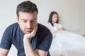 a man looking disappointed sitting on a bed with a woman in the background