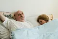 A mature couple lies in bed; the man stares at the ceiling with a sad expression while his wife sleep: Pain During Intercourse