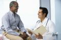 A doctor in a white coat holds a clipboard and talks to a patient sitting on an examination table. The patient, wearing glasses, a striped shirt, and khaki pants, smiles and listens attentively in a brightly lit medical office.