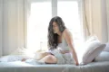 A woman in white sitting on her bed, holding her abdomen in pain for uti discharge in women