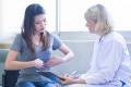 A young woman points to her abdomen while talking to a female doctor. The woman is wearing a gray shirt and jeans, and the doctor is in a white coat holding a clipboard. They are sitting in a well-lit room, engaging in a consultation.