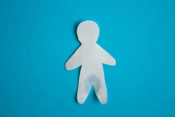 A cutout of a simple human figure made from white paper is placed against a blue background. The paper figure appears to have a small water stain around the lower abdomen area. The figure is simplistic with no facial details.