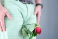 A person wearing light green patterned pants with a red rose tucked into the waistband. The person’s hands are placed on their hips, and they are wearing a black belt and a wristwatch. The background is plain gray, reminiscent of waiting room walls in clinics offering erectile dysfunction treatments near me.