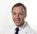 A man with short brown hair wearing a white medical coat and a dark tie is smiling at the camera. He is part of our team at Comprehensive Urology Los Angeles, known for having some of the best urologists in Los Angeles. The background is plain white.