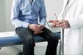 A person in a light blue shirt and dark jeans sits on a medical examination table, clasping their hands. A doctor in a white coat stands nearby holding a pair of glasses, engaged in conversation. Both individuals are only partially visible from the chest down.