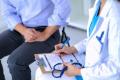 A medical professional in a white coat is seated and writing on a clipboard with a stethoscope placed on their lap. In front of the professional, a person is sitting with hands clasped. The setting appears to be a consultation or examination room.