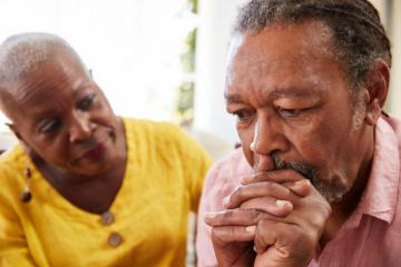 A concerned older woman with short hair and wearing a yellow top sits next to an older man with braided hair, a mustache, and a pink shirt, who looks distressed and is resting his chin on his hands, deep in thought. They are in a brightly lit room.