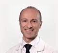 A man with a receding hairline, smiling and looking directly at the camera. He is wearing a white lab coat and a dark tie against a plain, light background. He represents our team at Comprehensive Urology Los Angeles, known for being among the best urologists in the area.