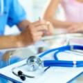 A close-up of a stethoscope resting on a clipboard with medical documents. In the blurred background, a doctor wearing a blue uniform and a patient wearing a pink top can be seen sitting at a table, engaged in a discussion.