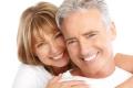 A smiling older couple with gray and blonde hair. The woman, embracing the man from behind, both dressed in white shirts, look happy and content against a white background. Their smiles radiate the benefits of adult circumcision and a life lived to the fullest.