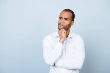 A man with short hair, dressed in a white button-down shirt, stands against a light gray background. He is looking thoughtfully into the distance with his hand resting on his chin and his other arm crossed over his chest.