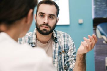 A bearded man in a plaid shirt gestures with his hand while speaking to another person in a room with blue walls. The other person's face is not visible, and blinds cover the windows in the background.
