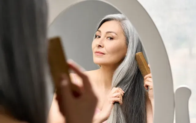 a woman with grey and black hair combs her hair while looking into a mirror