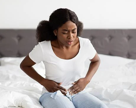 a young black lady sits on bed wearing white shirt and light blue jeans in visible pain